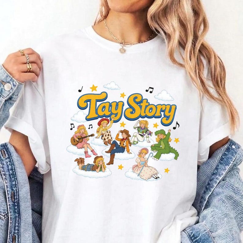 Pu&ograve; includere: Una t-shirt bianca con una stampa grafica colorata con il testo Tay Story in lettere gialle. Il design include diversi personaggi illustrati con capelli biondi seduti su nuvole, circondati da stelle gialle e note musicali.