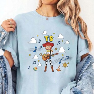 May include: A light blue t-shirt featuring a graphic of a cartoon cowgirl playing an acoustic guitar. The design includes the text TS, white clouds, musical notes, yellow stars, a colorful ball, a sheriff star badge, and an orange traffic cone.