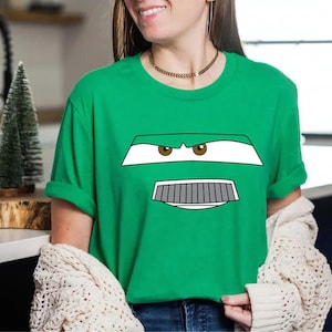 May include: Green t-shirt featuring a cartoon car face design with white, gray, and brown details. The design includes eyes, a grill, and a bumper. The shirt is a solid green color.
