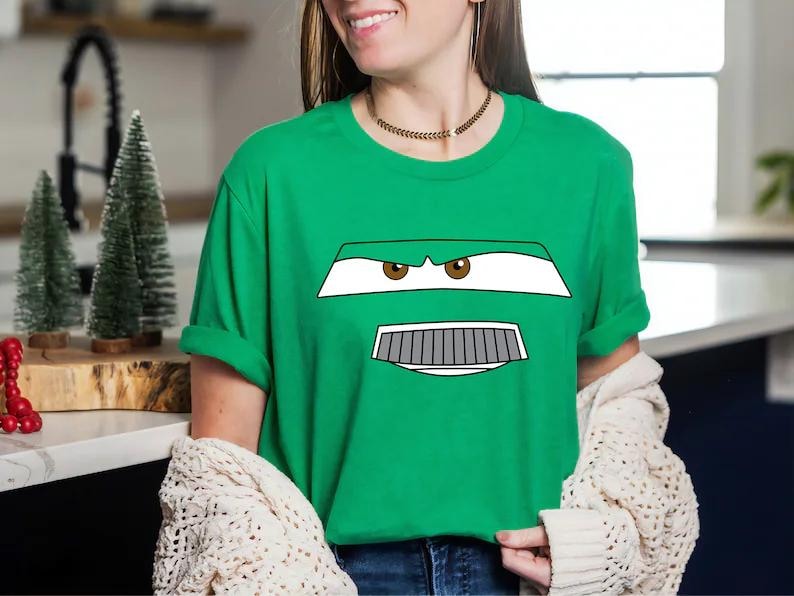 May include: Green t-shirt featuring a cartoon car face design with white, gray, and brown details. The design includes eyes, a grill, and a bumper. The shirt is a solid green color.