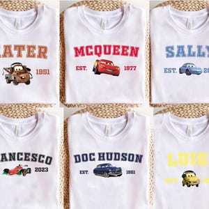 May include: White t-shirts with designs from the film Cars. Each shirt displays a character's name, such as "Mater" or "McQueen," alongside a car graphic and the year of establishment. The designs are in various colours.