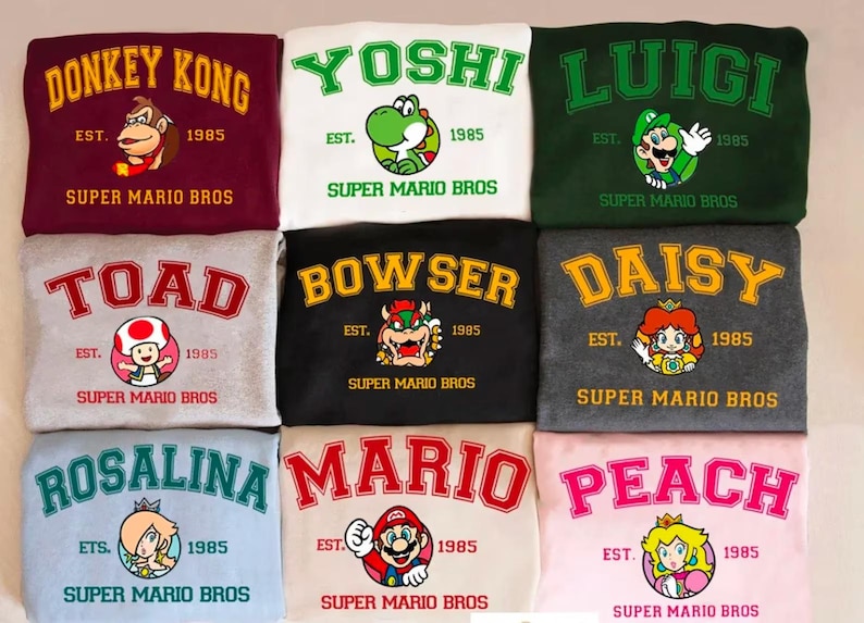 May include: Nine sweatshirts featuring Super Mario Bros characters. Each sweatshirt displays a character's name in a collegiate font, along with the text "EST. 1985" and "SUPER MARIO BROS." Characters include Donkey Kong, Yoshi, Luigi, Toad, Bowser, Daisy, Rosalina, Mario, and Peach.