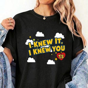 May include: A black short-sleeved t-shirt featuring a graphic print with white clouds, small yellow and green stars, and the text "I KNEW IT, I KNEW YOU" in yellow with a blue outline, plus a red heart containing the letters "TS" in yellow.