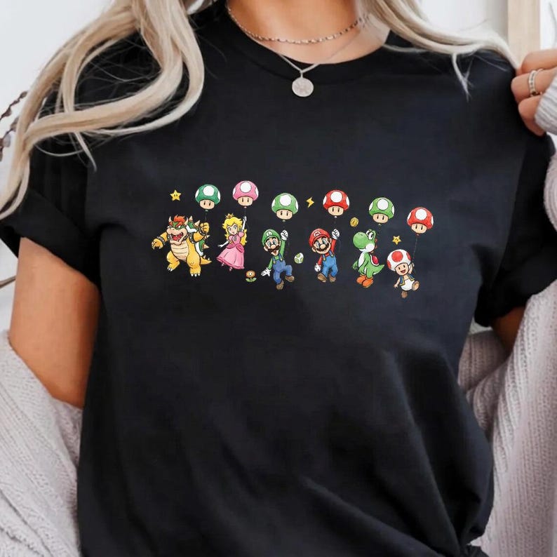 May include: A black short-sleeved t-shirt featuring a graphic print of various video game characters including Bowser, Princess Peach, Luigi, Mario, Yoshi, and Toad. Each character is holding a colorful mushroom-shaped balloon on a string.