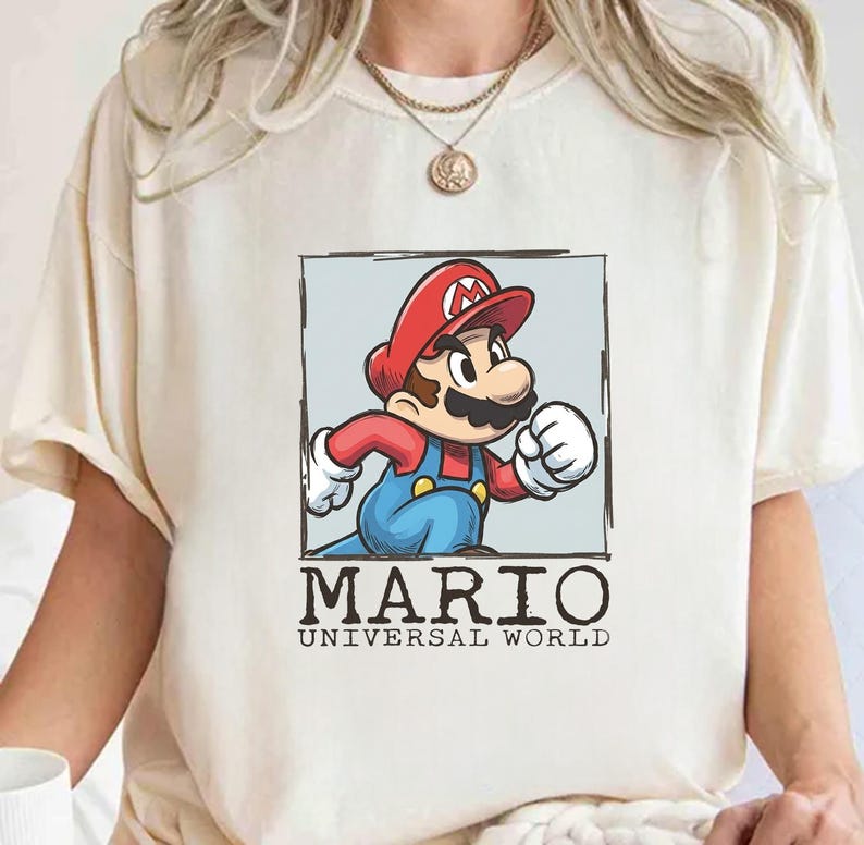 May include: A cream-colored t-shirt featuring a graphic print of Mario in a red cap and blue overalls inside a square frame. Below the frame, the text reads MARIO UNIVERSAL WORLD in black capital letters. The shirt has short sleeves and a crew neck.