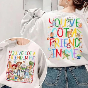 May include: White sweatshirt featuring a colorful graphic of characters and the phrase "YOU'VE GOT A FRIEND IN ME." The design includes cartoon figures and text in various colors. The back of the sweatshirt also has the same design.