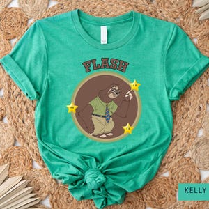 May include: Kelly green t-shirt featuring a cartoon sloth character wearing a green shirt and tie, with the word "FLASH" above the sloth. The sloth is inside a brown circle with three yellow stars.