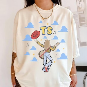 May include: An off-white t-shirt featuring a graphic of a person with blonde hair wearing a yellow plaid shirt and cow-print pants, holding a guitar and a red hat. The design includes blue clouds, yellow stars, musical notes, and the letters TS.
