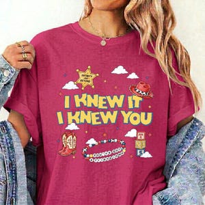 Puede incluir: Una camiseta magenta de manga corta con el texto amarillo I KNEW IT I KNEW YOU. El gr&aacute;fico incluye una estrella de sheriff dorada, un sombrero de vaquero rojo, un par de botas vaqueras rojas, una pulsera de cuentas y bloques de letras.