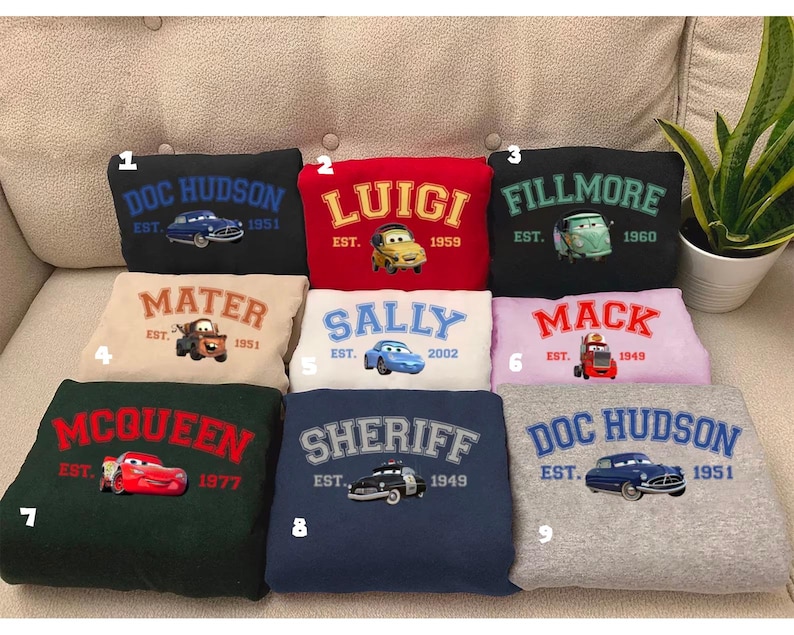 May include: Nine sweatshirts with embroidered cartoon car designs and names. The sweatshirts are in various colours: black, red, green, beige, white, pink, navy, and grey. Each sweatshirt features a different name: "Doc Hudson", "Luigi", "Fillmore", "Mater", "Sally", "Mack", "McQueen", "Sheriff", and "Doc Hudson".