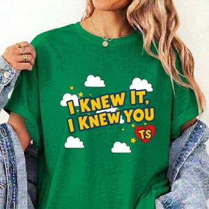 May include: A green t-shirt featuring the yellow text I KNEW IT, I KNEW YOU TS with a red heart, white clouds, and small yellow stars. The shirt has short sleeves and a crew neckline, layered under a blue denim jacket with silver buttons.