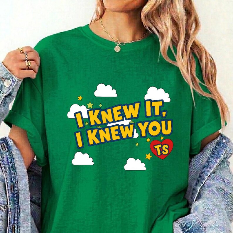 May include: A green t-shirt featuring the yellow text I KNEW IT, I KNEW YOU TS with a red heart, white clouds, and small yellow stars. The shirt has short sleeves and a crew neckline, layered under a blue denim jacket with silver buttons.