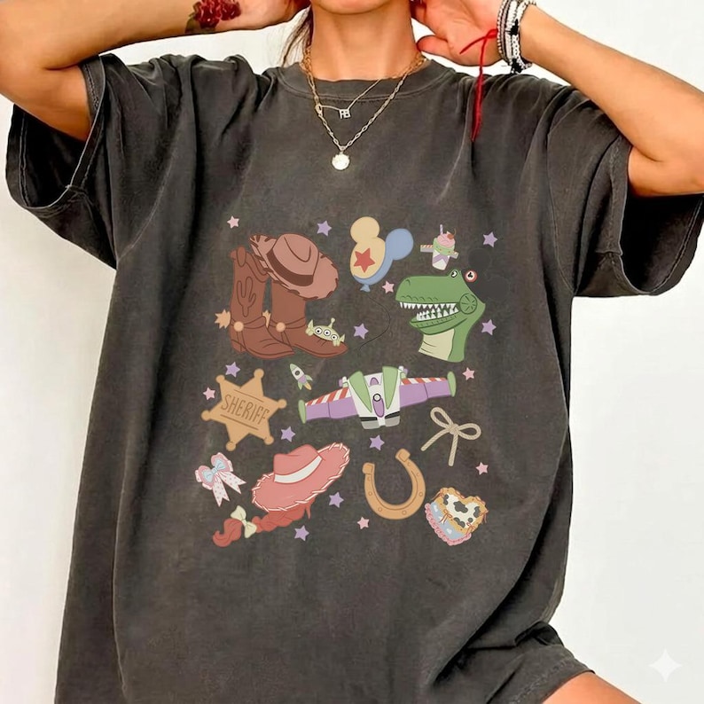 May include: Dark gray t-shirt featuring a colorful graphic of cartoon characters and symbols, including a cowboy hat, boots, a sheriff badge, and a dinosaur. The design also includes a rocket ship, a horseshoe, and a bow.