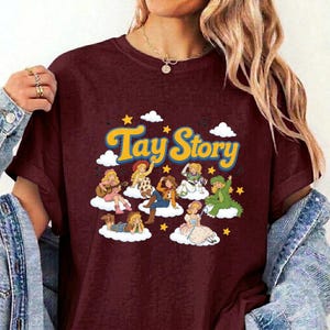 Pu&ograve; includere: Una t-shirt a maniche corte color bordeaux con una stampa grafica che riporta il testo "Tay Story" in lettere gialle. Il design include diversi personaggi dei cartoni animati seduti su nuvole bianche circondate da stelle gialle.