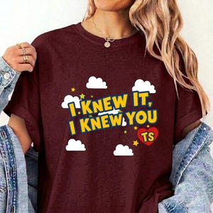 May include: A maroon short-sleeved t-shirt featuring a graphic print with white clouds, yellow stars, and a red heart containing the letters TS. The text reads I KNEW IT, I KNEW YOU in bold yellow letters with a dark blue outline against the maroon fabric.
