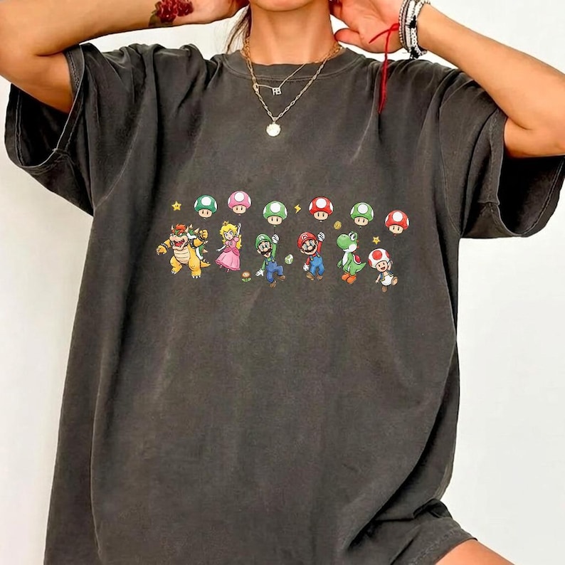 May include: A charcoal gray short-sleeve t-shirt featuring a graphic print of various Super Mario characters including Bowser, Princess Peach, Luigi, Mario, Yoshi, and Toad, interspersed with colorful mushrooms and small yellow stars.