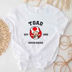 May include: White t-shirt with a black graphic of Toad from Super Mario Bros. The graphic includes the text "TOAD EST 1985 SUPER MARIO".
