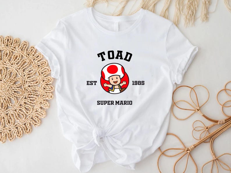 May include: White t-shirt with a black graphic of Toad from Super Mario Bros. The graphic includes the text "TOAD EST 1985 SUPER MARIO".