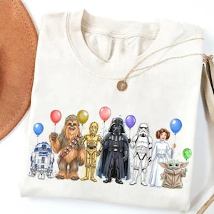 K&ouml;nnte beinhalten: Cremefarbenes T-Shirt mit einer farbenfrohen Illustration von Star Wars-Charakteren, die Ballons halten. Die Charaktere sind R2-D2, Chewbacca, C-3PO, Darth Vader, ein Sturmtruppler, Prinzessin Leia und Grogu.