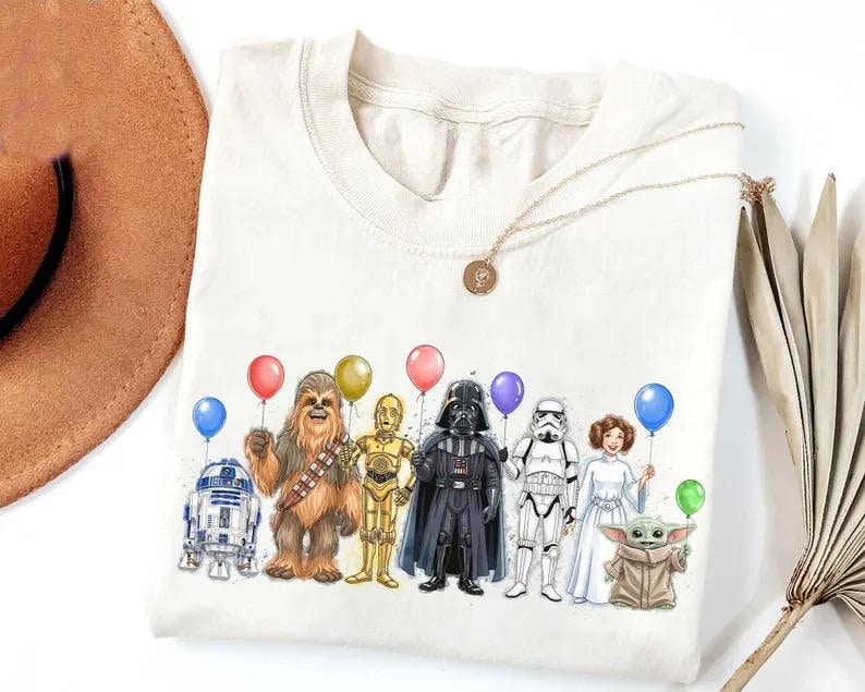 K&ouml;nnte beinhalten: Cremefarbenes T-Shirt mit einer farbenfrohen Illustration von Star Wars-Charakteren, die Ballons halten. Die Charaktere sind R2-D2, Chewbacca, C-3PO, Darth Vader, ein Sturmtruppler, Prinzessin Leia und Grogu.