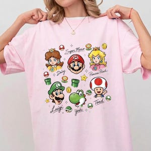 May include: A light pink t-shirt featuring a colorful graphic print of Super Mario characters including Mario, Luigi, Princess Peach, Daisy, Yoshi, and Toad, along with various game icons and the names of the characters written in cursive script.
