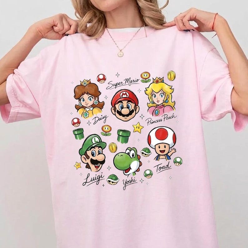 May include: A light pink t-shirt featuring a colorful graphic print of Super Mario characters including Mario, Luigi, Princess Peach, Daisy, Yoshi, and Toad, along with various game icons and the names of the characters written in cursive script.
