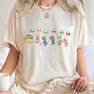 May include: A cream-colored short-sleeve t-shirt featuring a graphic print of various Nintendo characters including Bowser, Princess Peach, Luigi, Mario, Yoshi, and Toad, each holding a colorful balloon. The shirt has a crew neckline.