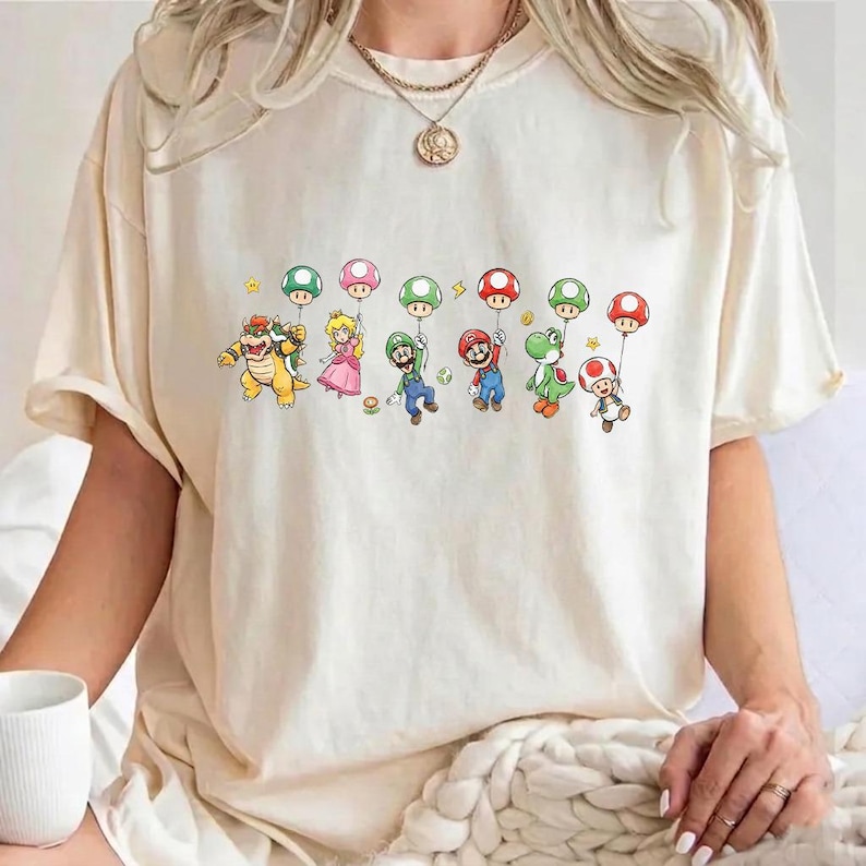 May include: A cream-colored short-sleeve t-shirt featuring a graphic print of various Nintendo characters including Bowser, Princess Peach, Luigi, Mario, Yoshi, and Toad, each holding a colorful balloon. The shirt has a crew neckline.