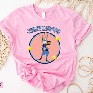 May include: Pink t-shirt featuring a cartoon image of Judy Hopps from Zootopia. The design includes the text "JUDY HOPPS" above a circular image of the character, with yellow star accents. The shirt is made of a soft material.