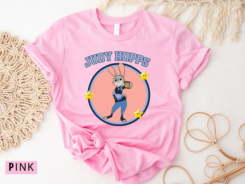 May include: Pink t-shirt featuring a cartoon image of Judy Hopps from Zootopia. The design includes the text "JUDY HOPPS" above a circular image of the character, with yellow star accents. The shirt is made of a soft material.
