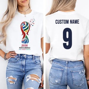 May include: White t-shirt featuring a colorful graphic with the text "2026 NORTH AMERICA USA CANADA MEXICO". The back of the shirt displays "CUSTOM NAME" above the number "9" in navy blue. The shirt is worn with blue jeans.