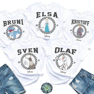 May include: White t-shirts featuring characters from the movie Frozen. Each shirt has a character's name in black block letters above a circular design with a checkered border. Characters include Bruni, Elsa, Kristoff, Sven, and Olaf.