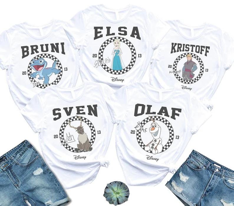 May include: White t-shirts featuring characters from the movie Frozen. Each shirt has a character's name in black block letters above a circular design with a checkered border. Characters include Bruni, Elsa, Kristoff, Sven, and Olaf.