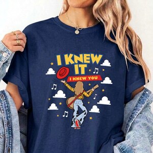 May include: A navy blue t-shirt featuring a graphic print of a person with long hair wearing a yellow plaid shirt and cow-print pants, holding a guitar. The text reads I KNEW IT I KNEW YOU, surrounded by white clouds, yellow stars, and music notes.