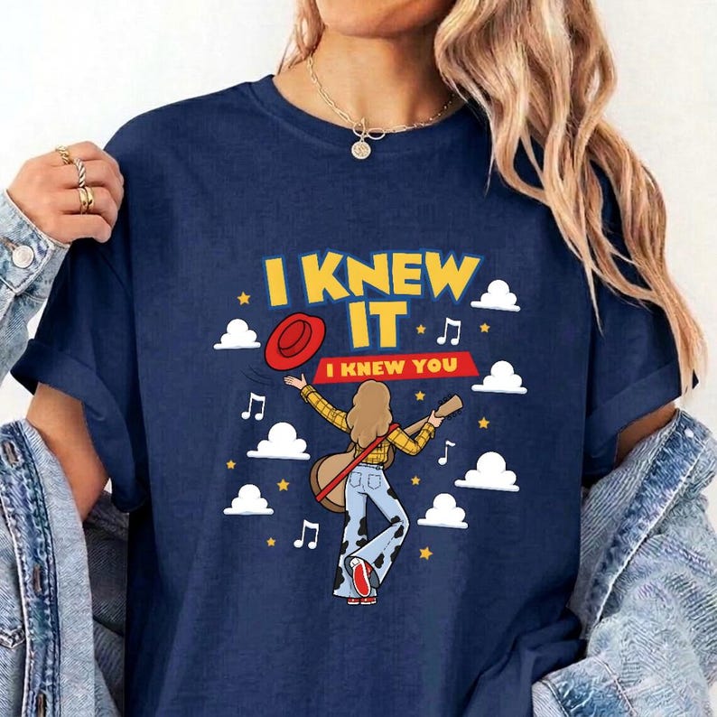 May include: A navy blue t-shirt featuring a graphic print of a person with long hair wearing a yellow plaid shirt and cow-print pants, holding a guitar. The text reads I KNEW IT I KNEW YOU, surrounded by white clouds, yellow stars, and music notes.