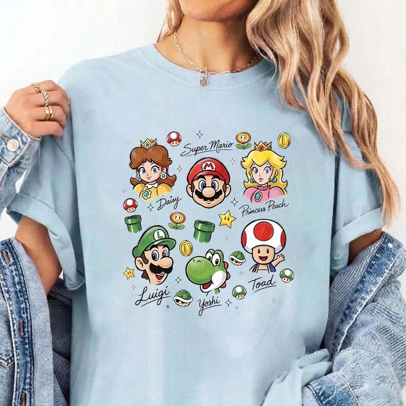 May include: A light blue t-shirt featuring a graphic print of Super Mario characters including Mario, Luigi, Princess Peach, Daisy, Yoshi, and Toad. The design includes various game icons like mushrooms, coins, stars, and pipes with handwritten names.