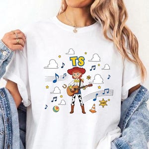 May include: A white t-shirt featuring a graphic of a cartoon cowgirl playing an acoustic guitar. The design includes the text TS, musical notes, clouds, stars, a ball, and a traffic cone. The cowgirl wears a red hat, yellow shirt, and cow-print pants.