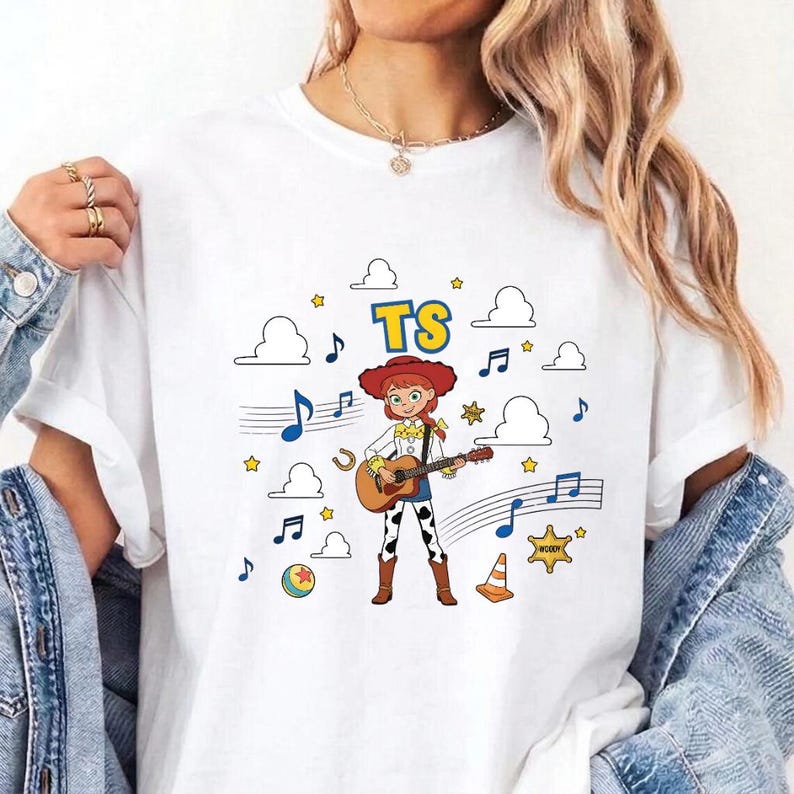 May include: A white t-shirt featuring a graphic of a cartoon cowgirl playing an acoustic guitar. The design includes the text TS, musical notes, clouds, stars, a ball, and a traffic cone. The cowgirl wears a red hat, yellow shirt, and cow-print pants.