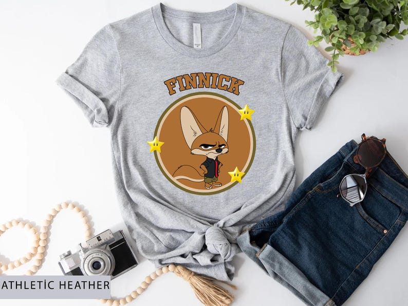 May include: Athletic heather gray t-shirt featuring a cartoon image of Finnick from Zootopia. The design includes Finnick's image inside a brown circle with the name "Finnick" above and yellow stars on each side.