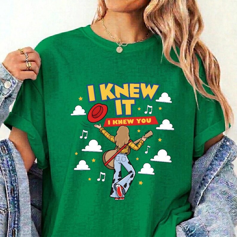 May include: A green short-sleeve t-shirt featuring a graphic of a person with long hair wearing a yellow plaid shirt and cow-print pants, holding a guitar. The text reads I KNEW IT and I KNEW YOU, surrounded by white clouds, stars, and music notes.
