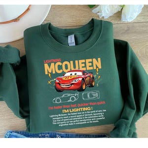 May include: A dark green sweatshirt featuring Lightning McQueen, a red race car from the Cars movie. The design includes the text "LIGHTNING MCQUEEN" and "I'm faster than fast. Quicker than quick. I'M LIGHTING!"
