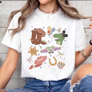 May include: White t-shirt featuring a colorful graphic design with cowboy boots, a sheriff badge, a green dinosaur, a space ship, and other cartoon elements. The design includes stars, a horseshoe, and a pink hat.