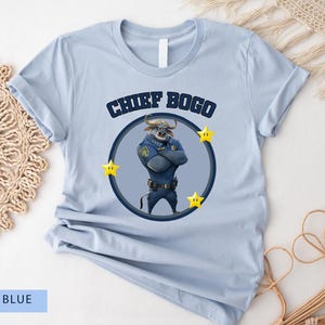 May include: Light blue t-shirt featuring a cartoon image of Chief Bogo from Zootopia. The design includes the text "CHIEF BOGO" above a circular image of the character, surrounded by yellow stars. The shirt is made of a soft material.
