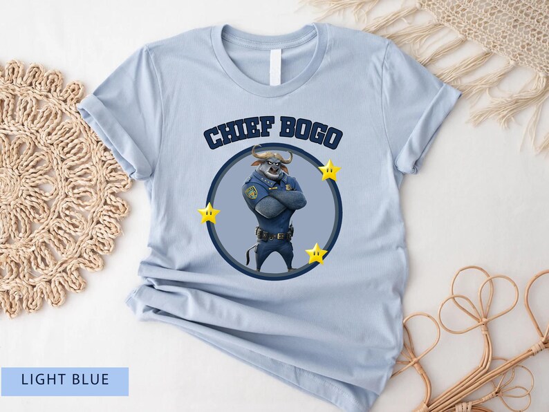 May include: Light blue t-shirt featuring a cartoon image of Chief Bogo from Zootopia. The design includes the text "CHIEF BOGO" above a circular image of the character, surrounded by yellow stars. The shirt is made of a soft material.