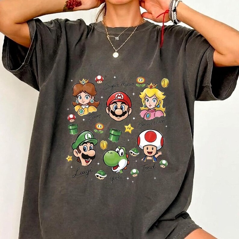 May include: A charcoal gray short-sleeve t-shirt featuring a graphic print of Super Mario characters including Mario, Luigi, Princess Peach, Princess Daisy, Yoshi, and Toad, along with various game icons like coins, stars, and pipes.