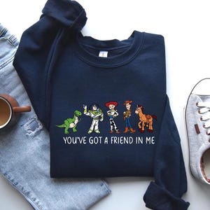 May include: Navy blue sweatshirt featuring a graphic of cartoon characters with the text "YOU'VE GOT A FRIEND IN ME". The design includes a green dinosaur, a white and green astronaut, a cowgirl, a cowboy, and a horse.