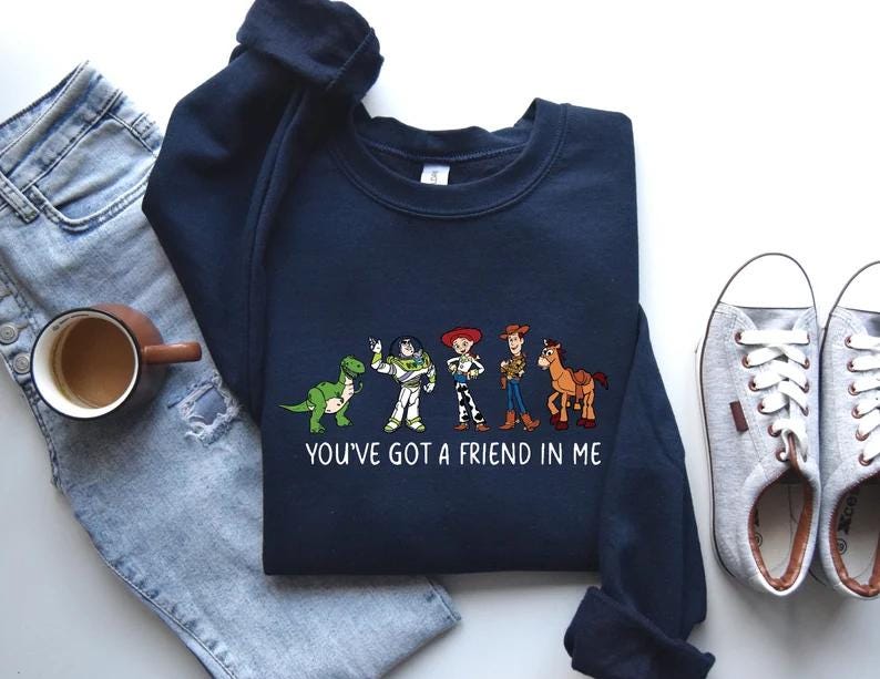 May include: Navy blue sweatshirt featuring a graphic of cartoon characters with the text "YOU'VE GOT A FRIEND IN ME". The design includes a green dinosaur, a white and green astronaut, a cowgirl, a cowboy, and a horse.