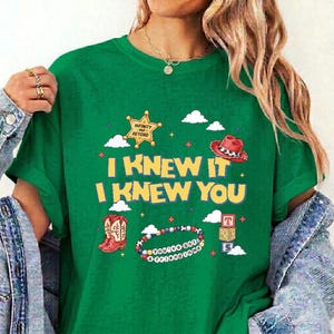 Op de afbeelding: Een groen t-shirt met een grafische print met de tekst I KNEW IT I KNEW YOU, een gele sheriffster, een rode cowboyhoed, een paar rode cowboylaarzen, een kralenarmband met de tekst YOU'VE GOT A FRIEND IN ME en gestapelde blokken.