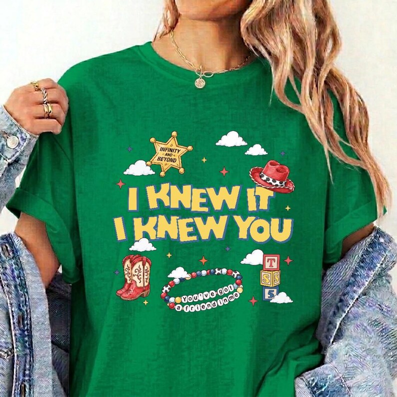 Op de afbeelding: Een groen t-shirt met een grafische print met de tekst I KNEW IT I KNEW YOU, een gele sheriffster, een rode cowboyhoed, een paar rode cowboylaarzen, een kralenarmband met de tekst YOU'VE GOT A FRIEND IN ME en gestapelde blokken.