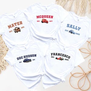 May include: White t-shirts with character names and car graphics from the film Cars. Each shirt has a character's name, a car image, and the year of their introduction. Characters include Mater, McQueen, Sally, Doc Hudson, and Francesco.